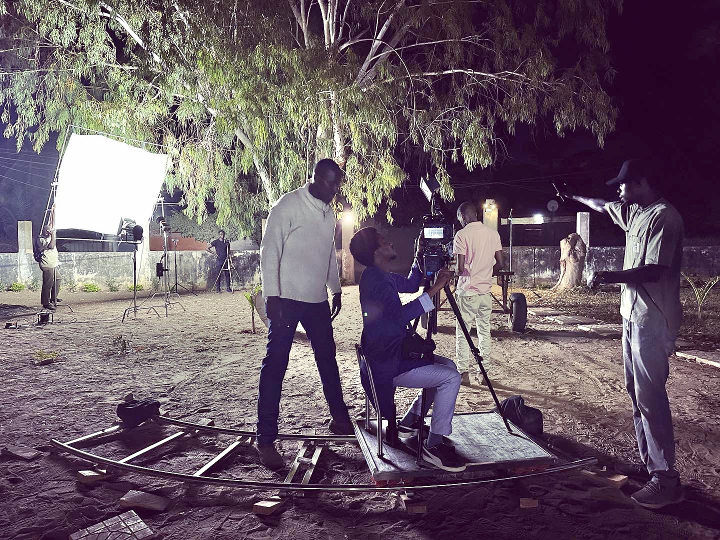 Filmmakers setting up a camera on a film set at night, with lighting equipment and crew members in the background